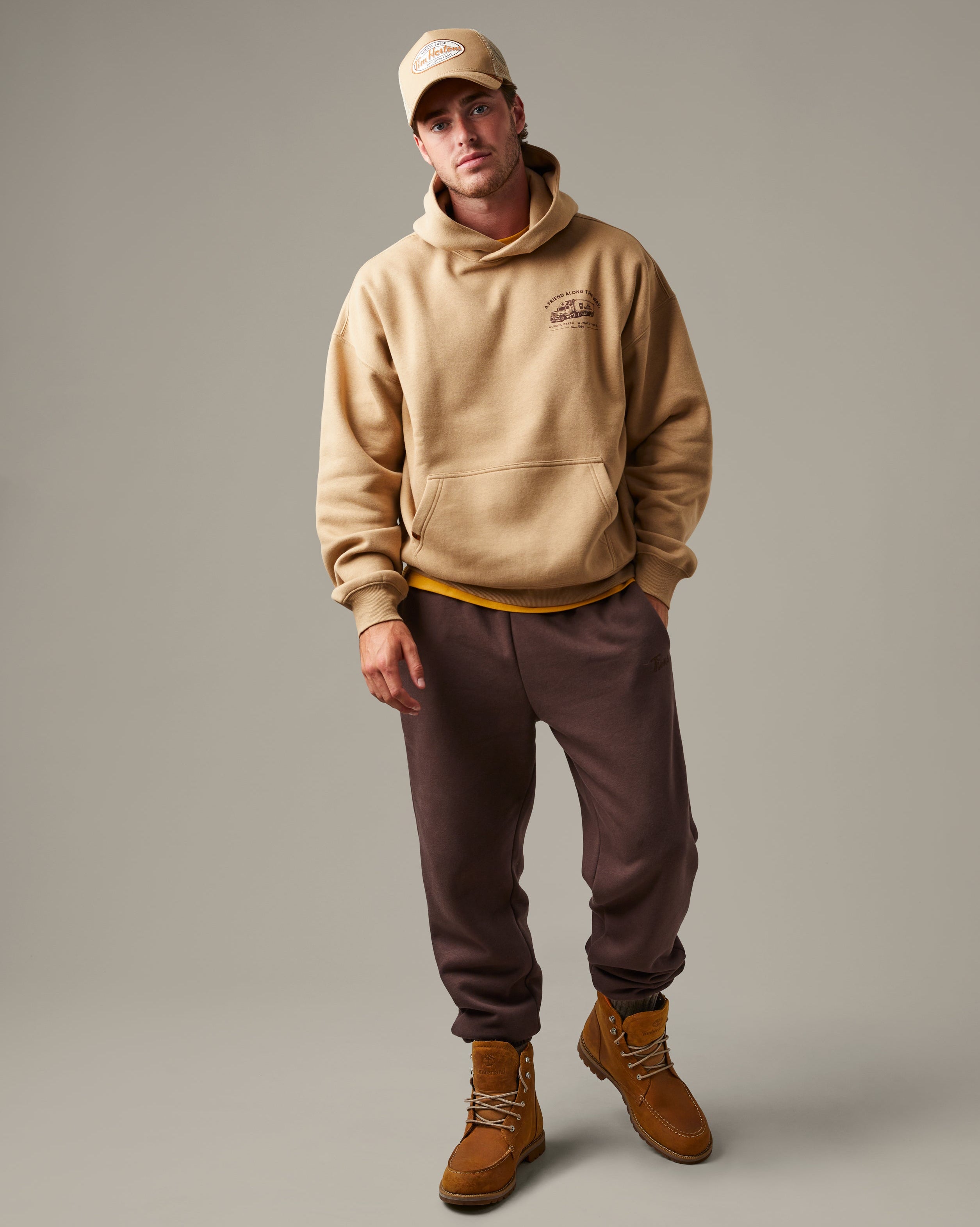 Beige Tim Hortons pullover hoodie with a kangaroo pocket and small A Friend Along the Way delivery truck graphic on the chest. | Chandail à capuchon Tim Hortons beige avec poche kangourou et petite illustration de camion de livraison « Un ami sur la route » sur la poitrine. - Image #1