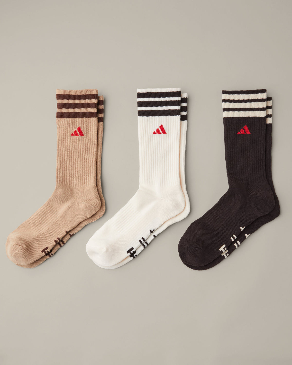 Three pairs of men’s adidas® crew socks in tan, cream, and espresso brown with striped cuffs and red adidas® logos. - Image #1