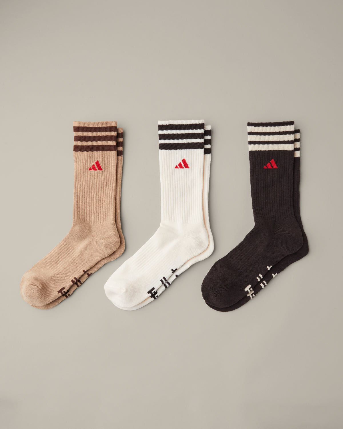 Three pairs of men’s adidas® crew socks in tan, cream, and espresso brown with striped cuffs and red adidas® logos. - Image #1